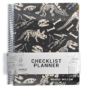 May include: A spiral-bound checklist planner with a black cover featuring a white dinosaur skeleton pattern. A white paper band wraps around the center, printed with the text CHECKLIST PLANNER, MANDY WILLOW, and various hand-drawn icons.