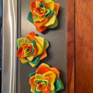 May include: Three colourful paper flower magnets in shades of green, yellow, and orange. The flowers are layered and have a 3D effect.