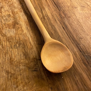 Può includere: Un cucchiaio di legno con un manico lungo e una ciotola arrotondata. Il cucchiaio è fatto di legno marrone chiaro e ha una finitura naturale.