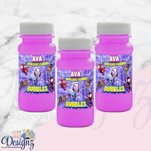 May include: Three bright pink plastic bubble bottles with white screw-on caps. Each bottle features a custom label with the text AVA, AMAZING FRIENDS, and BUBBLES, decorated with colourful cartoon superhero characters against a purple web background.