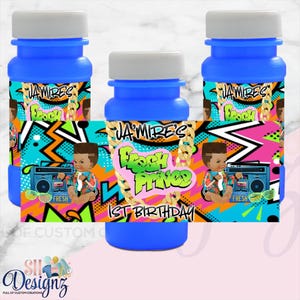 May include: Three blue plastic bubble solution bottles with custom labels featuring a cartoon baby, a boombox, and the text "JA'MIRE'S FRESH PRINCE 1ST BIRTHDAY" in bright graffiti-style lettering against a colorful, abstract geometric background.