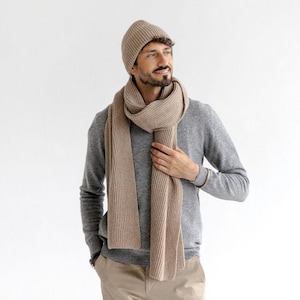 以下が含まれることがあります： ベージュのリブ編みニット帽とスカーフ、グレーのセーター、カーキ色のパンツを着用した人。スカーフは首に巻かれています。背景は無地の白です。