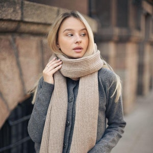 以下が含まれることがあります： グレーのカーディガンと白いパンツにベージュのニットスカーフを巻いた女性。スカーフは長く、リブ編みになっている。