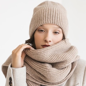 以下が含まれることがあります： ベージュのニット帽とマフラーを着用した女性。ベージュのコートを羽織っています。帽子とマフラーはリブ編みで、コートはボタン留めです。
