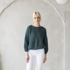 May include: A woman wearing a teal blue sweater and white trousers stands in front of a white wall with white paper lanterns hanging from the ceiling. She is wearing white heeled sandals.