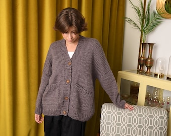 May include: A woman wearing a gray knitted cardigan with wooden buttons and two pockets. She is wearing black pants and leopard print shoes. She is standing in front of a yellow curtain and a chair with a green cushion.