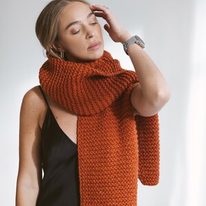 May include: A woman wearing a black slip dress with a chunky knit orange scarf. She is wearing a silver watch on her left wrist.