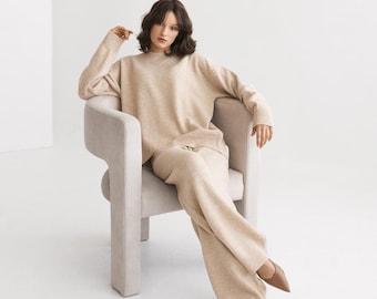 May include: A woman wearing a beige sweater and matching pants sits in a light beige armchair. She is looking at the camera.