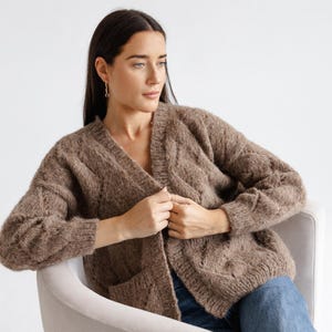 May include: A brown, open-front cardigan with a textured knit pattern. The sweater has a V-neck and two front pockets. The sleeves are long, and the cardigan is worn over blue jeans. The model is seated in a light-coloured chair.