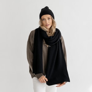 以下が含まれることがあります： 黒いニット帽とスカーフのセット。スカーフは長く長方形で、帽子はぴったりとしたキャップです。セットはリブ編みの素材で作られています。画像内の人物は、茶色のセーターと白いパンツを着用しています。