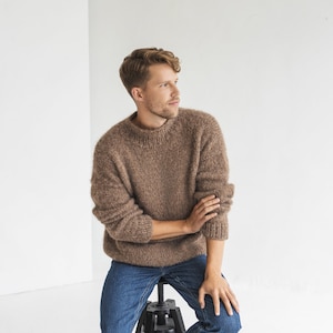 May include: A man wearing a brown fuzzy sweater and blue jeans sits on a black stool. He is looking to the right of the frame.