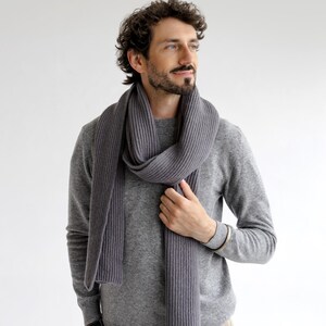 May include: A gray ribbed scarf draped over a gray sweater. The scarf is made of a soft material and has a vertical ribbed design. The person is wearing a gray sweater and khaki pants. The background is plain white.
