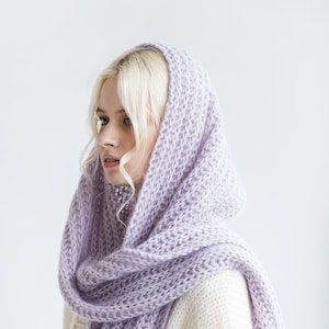 May include: A woman wearing a light purple knitted scarf with a hood. The scarf is draped around her neck and head, and she is looking to the side.