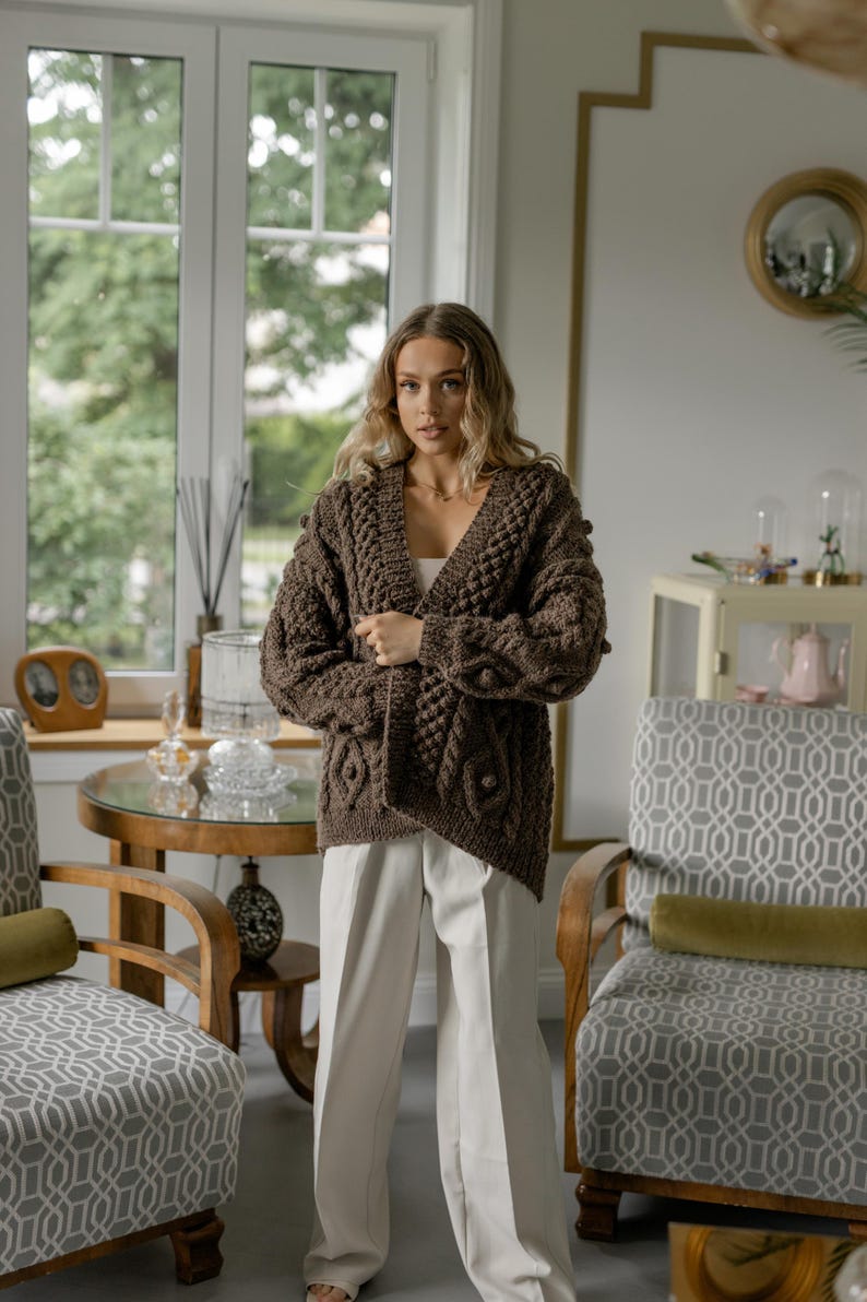 May include: A brown, chunky knit cardigan with a V-neck and intricate cable patterns. The long-sleeved sweater features textured bobbles and a relaxed fit. The model is wearing the cardigan with white pants. The background includes a window, chairs, and a table.