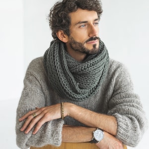 Peut inclure: Un homme portant un pull en laine gris et une écharpe en laine vert foncé. Il est assis sur une chaise en bois et regarde vers la droite. Il porte une montre au poignet gauche et une bague à la main droite.