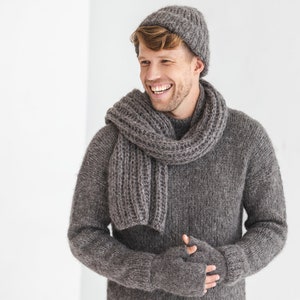 Puede incluir: Un hombre con un suéter, una bufanda y un gorro de punto grises. Está sonriendo y mirando hacia un lado.