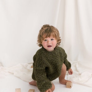 May include: A young child with curly brown hair wearing a green knitted sweater plays with wooden blocks on a white surface.