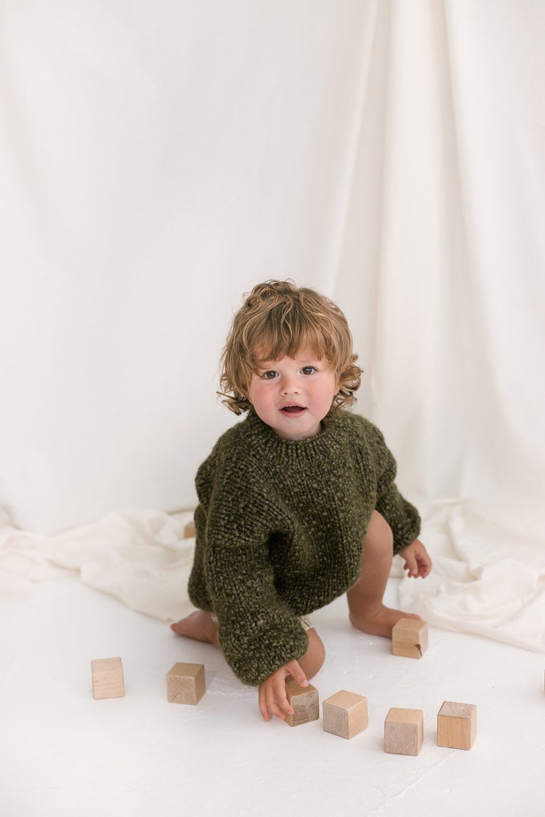 May include: A young child with curly brown hair wearing a green knitted sweater plays with wooden blocks on a white surface.