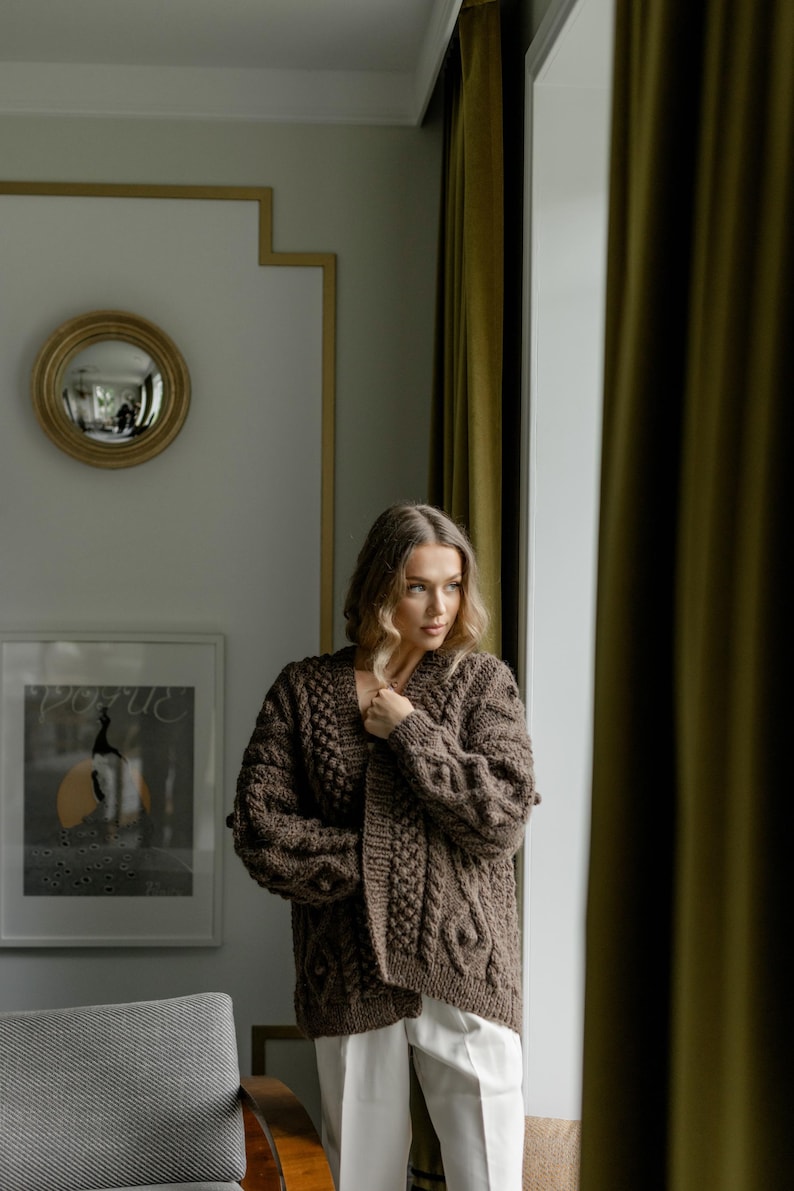 May include: A woman wearing a brown cable-knit cardigan with a textured pattern. The open-front sweater has a relaxed fit and is paired with white pants. The room features a gold-framed mirror and a framed Vogue print.
