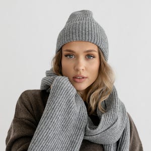 以下が含まれることがあります： グレーのリブ編みニット帽とスカーフのセット。スカーフは首と肩に巻かれています。モデルは茶色のセーターを着ています。背景は白です。