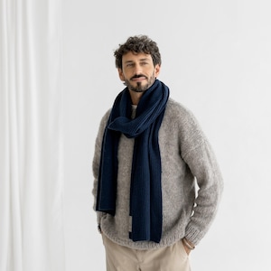 以下が含まれることがあります： ネイビーブルーのリブ編みスカーフとグレーのセーターを着た男性。スカーフは首に巻き付けられています。男性はベージュのパンツも着用しています。背景は白い壁です。