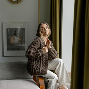 May include: A brown, chunky-knit cardigan with a cable-knit design and textured sleeves. The open-front sweater is worn with cream-colored pants. The image is set in a room with a sofa, curtains, and a framed print.