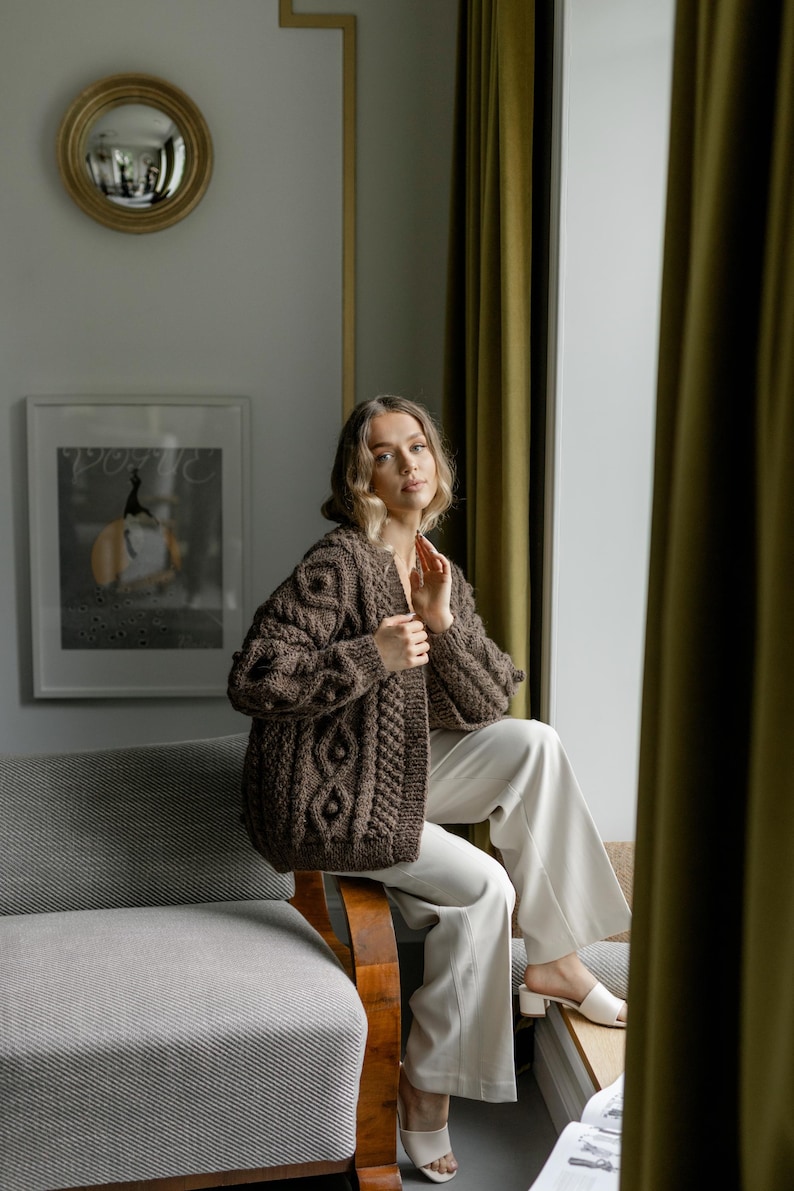 May include: A brown, chunky-knit cardigan with a cable-knit design and textured sleeves. The open-front sweater is worn with cream-colored pants. The image is set in a room with a sofa, curtains, and a framed print.