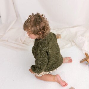 May include: A young child wearing a green knitted sweater sits on a white surface with wooden blocks scattered around them.