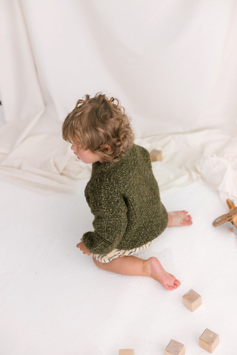 May include: A young child wearing a green knitted sweater sits on a white surface with wooden blocks scattered around them.