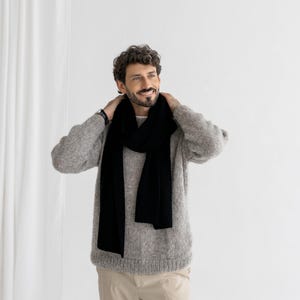 以下が含まれることがあります： グレーのセーターの上に黒いスカーフ。スカーフは長く長方形です。セーターは明るいグレーで、テクスチャード加工が施されています。ベージュのパンツを履いています。