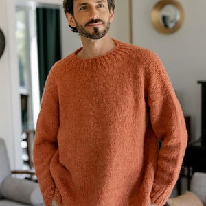 Pode incluir: Um suéter de malha cor ferrugem com gola redonda. O suéter tem mangas compridas e um corte folgado. A pessoa está usando calças cáqui. O fundo mostra uma sala de estar com uma paleta de cores neutras.