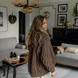May include: A brown cable-knit cardigan with a textured pattern. The sweater has a relaxed fit and long sleeves. The model is wearing white pants. The background includes a living room with a sofa, a coffee table, and a chandelier.