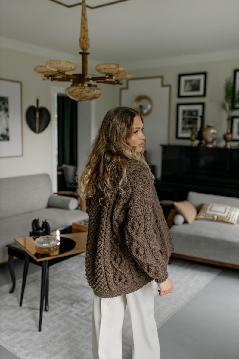 May include: A brown cable-knit cardigan with a textured pattern. The sweater has a relaxed fit and long sleeves. The model is wearing white pants. The background includes a living room with a sofa, a coffee table, and a chandelier.