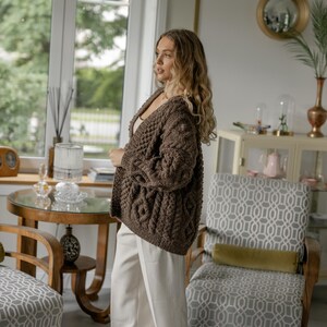 May include: A brown cable-knit cardigan with a V-neck. The open-front sweater has a textured pattern and is worn with white pants. The setting is a well-lit room with a window and decorative furniture.