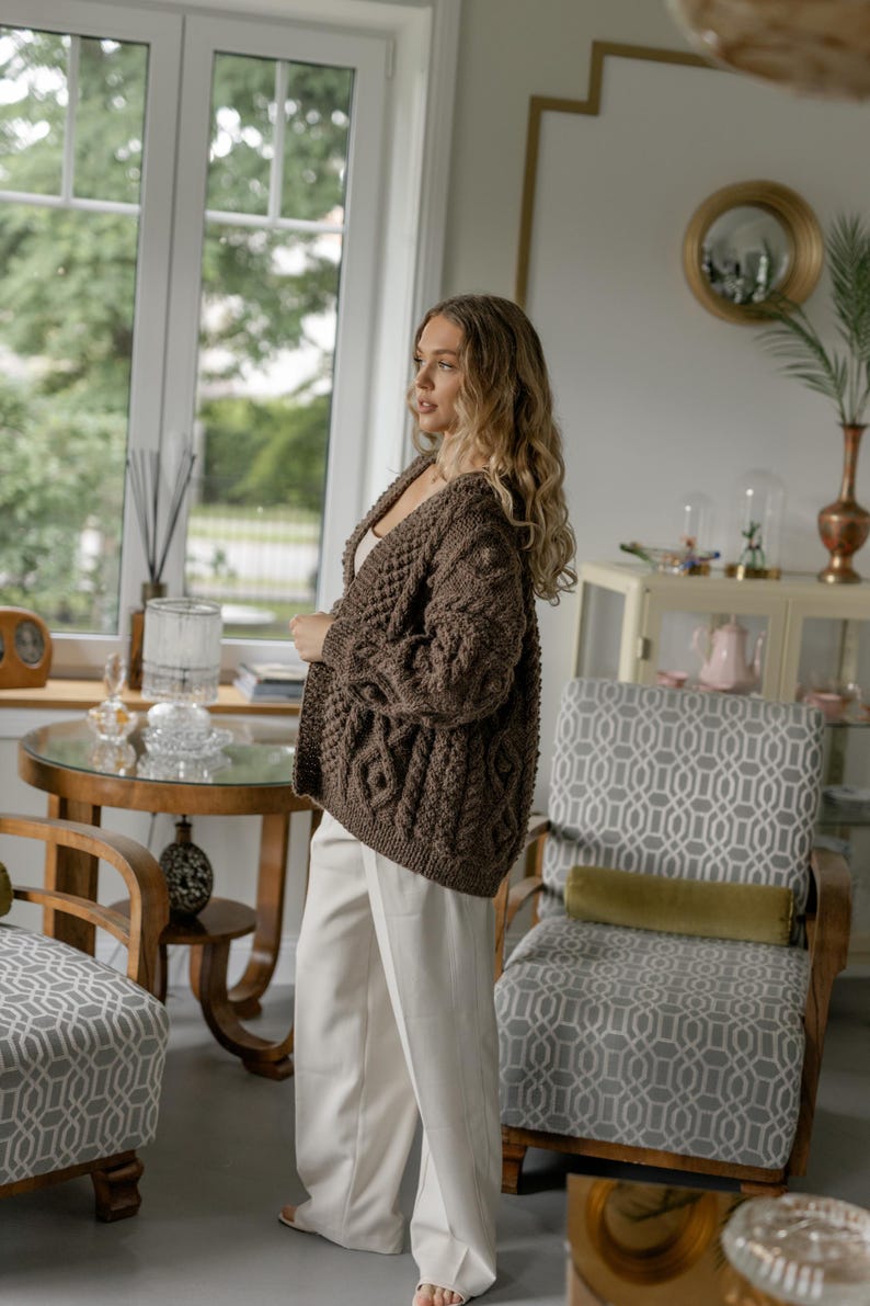May include: A brown cable-knit cardigan with a V-neck. The open-front sweater has a textured pattern and is worn with white pants. The setting is a well-lit room with a window and decorative furniture.