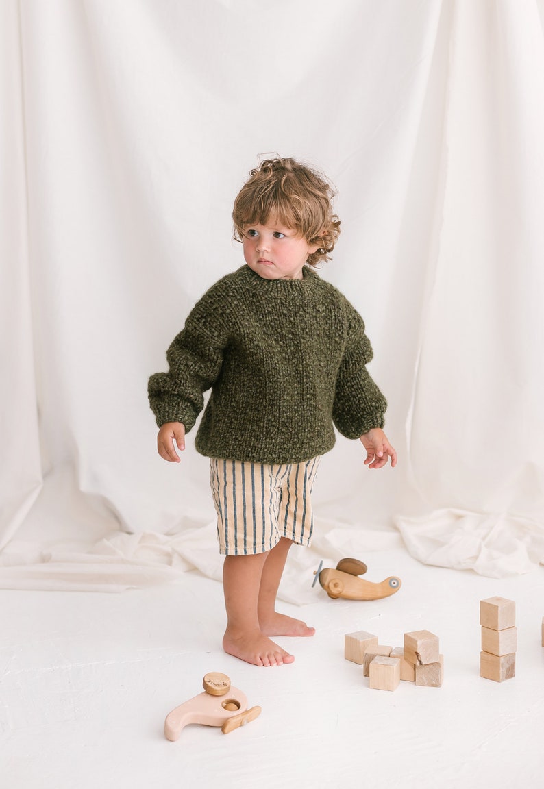 May include: A young child wearing a green knitted sweater and striped pants plays with wooden toys, including a helicopter and building blocks.