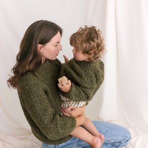 May include: A woman wearing a green sweater and blue jeans is holding a young child wearing a green sweater. The child is holding a small wooden block. They are both looking at each other.