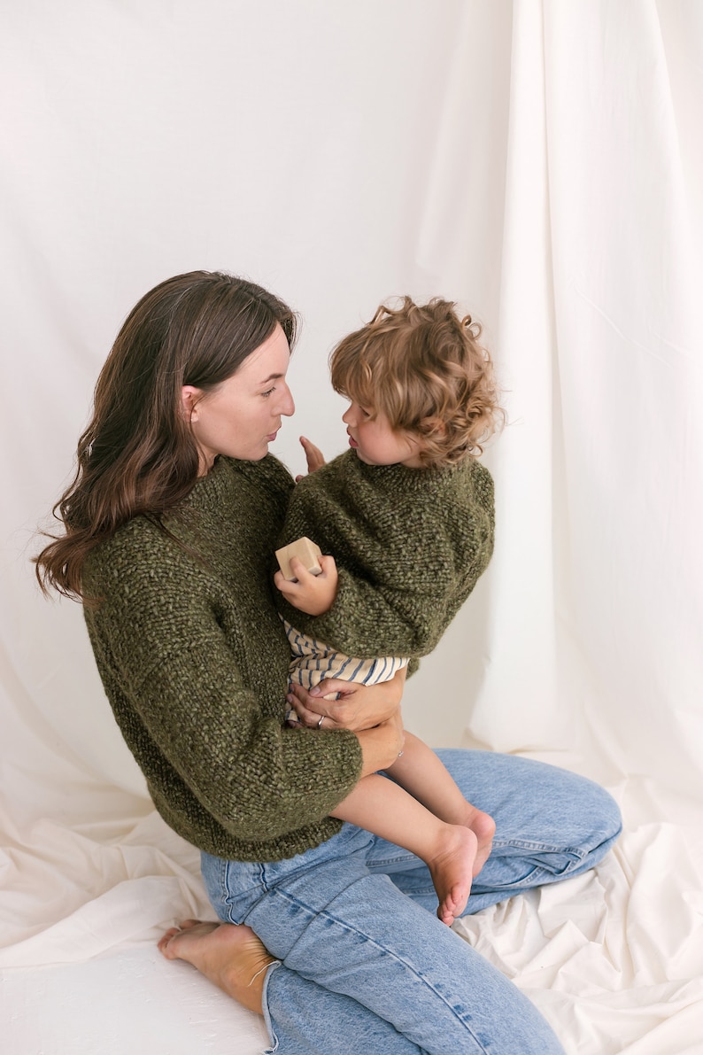 May include: A woman wearing a green sweater and blue jeans is holding a young child wearing a green sweater. The child is holding a small wooden block. They are both looking at each other.