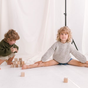 May include: Two children playing with wooden blocks. One child is wearing a green sweater and is building a tower of blocks. The other child is wearing a gray sweater and is sitting with their legs spread out.