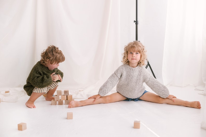 May include: Two children playing with wooden blocks. One child is wearing a green sweater and is building a tower of blocks. The other child is wearing a gray sweater and is sitting with their legs spread out.