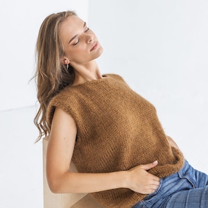 Könnte beinhalten: Eine Frau in einem braunen, ärmellosen Strickpullover und blauen Jeans sitzt auf einem hellen Holzhocker.