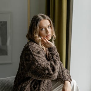 May include: A brown cable-knit cardigan with a textured pattern. The long-sleeved sweater has a relaxed fit and is worn with white pants. The cardigan is a warm, cozy garment, perfect for layering in cooler weather.