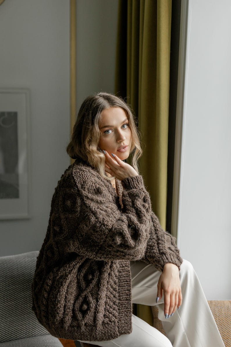 May include: A brown cable-knit cardigan with a textured pattern. The long-sleeved sweater has a relaxed fit and is worn with white pants. The cardigan is a warm, cozy garment, perfect for layering in cooler weather.