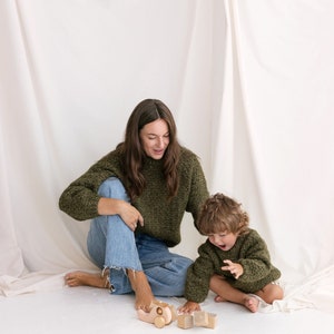 May include: A woman and a child are sitting on a white floor, playing with wooden toys. The woman is wearing a green sweater and blue jeans. The child is wearing a green sweater. The toys include a wooden car and blocks.