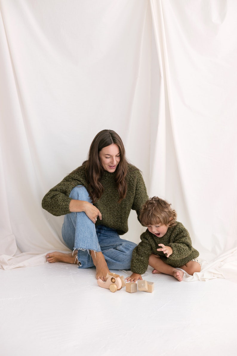 May include: A woman and a child are sitting on a white floor, playing with wooden toys. The woman is wearing a green sweater and blue jeans. The child is wearing a green sweater. The toys include a wooden car and blocks.