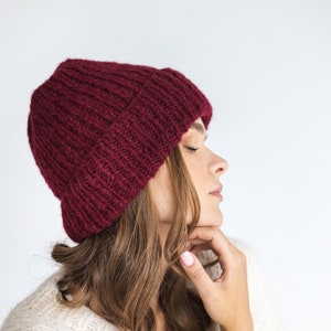 May include: A woman wearing a burgundy knitted beanie and a cream knitted sweater. Her hair is long and brown, and she is looking down.