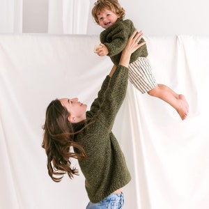 May include: A woman in a green sweater and blue jeans is holding a young child in the air. The child is wearing a green sweater and striped shorts. They are both smiling.