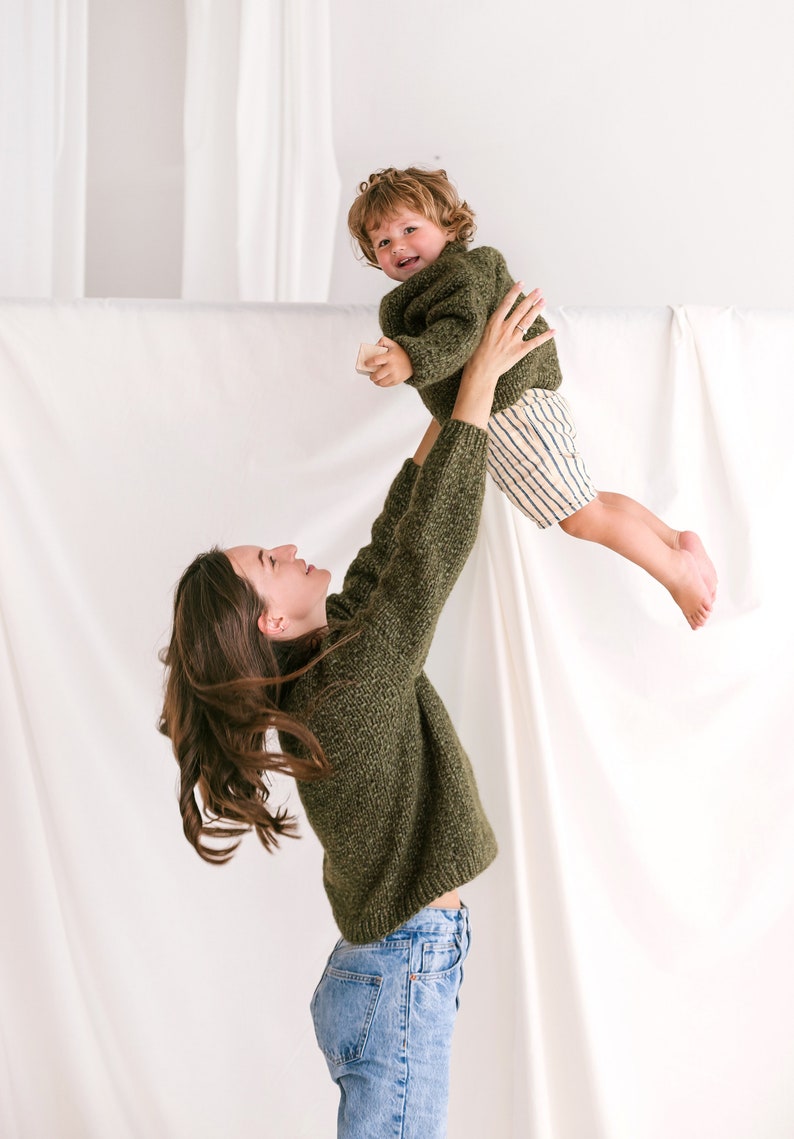 May include: A woman in a green sweater and blue jeans is holding a young child in the air. The child is wearing a green sweater and striped shorts. They are both smiling.