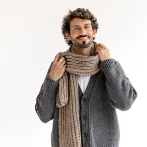 以下が含まれることがあります： ボタン付きのグレーのカーディガンとベージュのスカーフ。スカーフは明るい茶色で、首に巻かれています。カーディガンは白いシャツとベージュのパンツと組み合わせています。