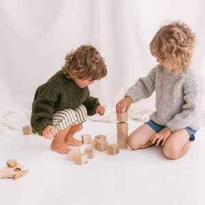 May include: Two children play with wooden blocks and a wooden toy helicopter. One child is wearing a green sweater and the other is wearing a gray sweater. The blocks are stacked in a tower.